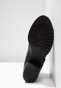 Bota de cuero negro con suela texturizada que presenta un patrón de clavos. El tacón es sólido y en forma de bloque, lo que demuestra una construcción duradera.