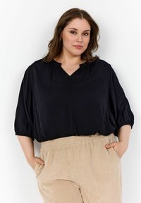Blusa nera con scollo a V, maniche 3/4 e polsini arricciati, abbinata a pantaloni beige a coste con vita elastica e tasche laterali.