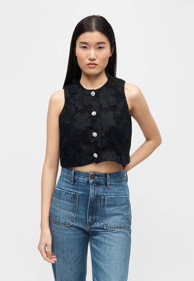 Νεαρή γυναίκα με μακριά σκούρα μαλλιά φοράει μαύρο αμάνικο κοντό τοπ με λουλουδάτη δαντέλα και διακοσμητικά κουμπιά, συνδυασμένο με ψηλόμεσα μπλε τζιν.