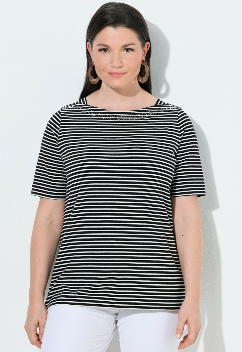 Kobieta z długimi ciemnymi włosami, nosząca duże brązowe kolczyki, czarno-białą koszulkę w paski z krótkim rękawem oraz białe spodnie, stojąca na jednolitym tle.