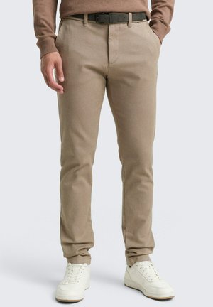 Man wearing beige slim-fit trousers, a brown sweater tucked in, black belt, and white sneakers, standing with hands partially in pockets.