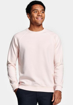 Jonge man die glimlacht, gekleed in een lichtroze trui met ronde hals en donkere broek, staand met ontspannen handen, witte achtergrond.