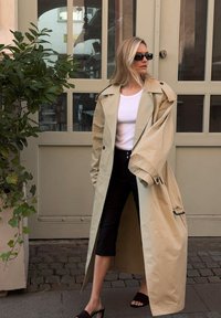 Beige trench coat with large collar, black buttons, and side pockets, worn over a white top and black cropped pants. Black high heels.
