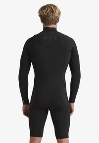 Ajuste completo de neopreno en negro, con mangas largas, cremallera trasera, costuras texturizadas y un logo en la parte superior de la espalda. Material suave y elástico.