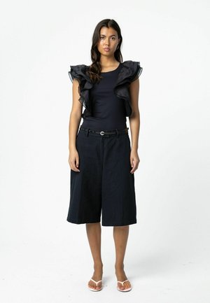 Vrouw met een mouwloze zwarte top met geplooide schouders, brede zwarte knielange shorts, een dunne zwarte riem en witte sandalen, staand tegen een witte achtergrond.