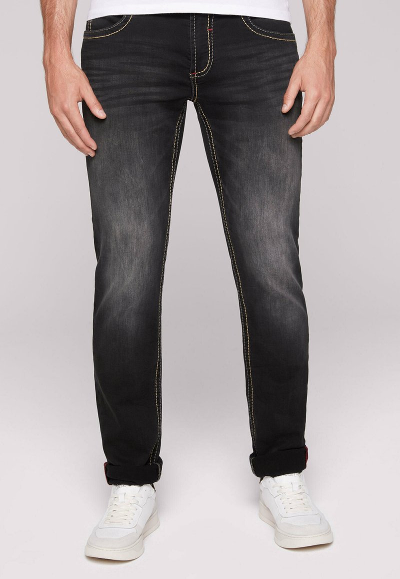 Schwarze Denim-Jeans mit leichter Verwaschung, gelben Nähten und schmaler Passform. Umschlagbare Säume. Kombiniert mit weißen Sneakern.