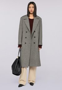 Donna in piedi che indossa un lungo cappotto grigio doppiopetto, pantaloni larghi beige, top bordeaux, scarpe nere a punta, tenendo una grande borsa nera.
