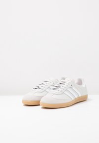 Zapatillas blancas con punteras de ante y laterales de cuero, que cuentan con tres rayas grises. Suela de goma con textura. Los cordones son de color gris claro.