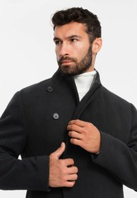 Abrigo de lana negro con cuello alto, cierre de doble botón y diseño estructurado. El forro interior presenta un sutil patrón de cuadros.