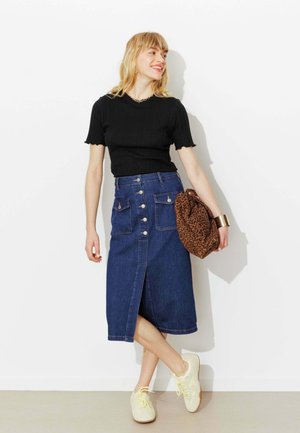 Femme souriante de profil portant un haut noir à manches courtes, une jupe midi en jean boutonnée, des baskets blanches, un sac à imprimé léopard et un bracelet en or.