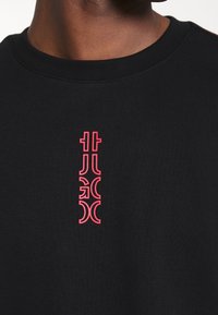 Gros plan sur une personne portant une chemise noire avec un design graphique rose vertical sur la poitrine.