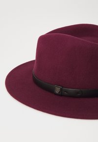 Chapeau en feutre bordeaux à large bord, orné d'un bandeau en cuir noir et d'un détail en métal.