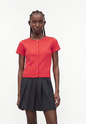 Vrouw met gevlochten haar, gekleed in een rood kort mouw t-shirt met knopen en een zwarte geplooide rok, staand tegen een effen witte achtergrond.