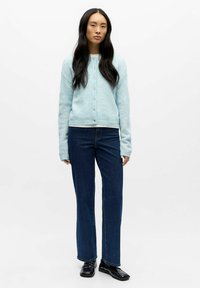 Lyseblå strikket cardigan med rund hals og knapper foran, kombinert med mørkeblå jeans med rette ben og blanke svarte sko.