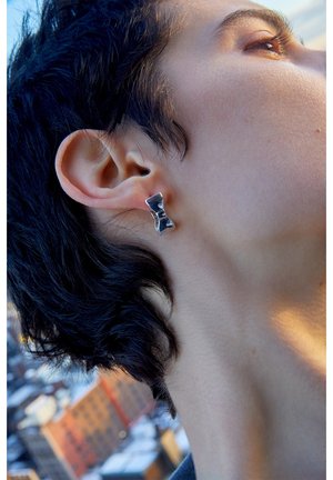 Gros plan d'une personne aux cheveux courts et foncés portant une boucle d'oreille argentée en forme de nœud papillon, la lumière du soleil illuminant sa joue et son cou.