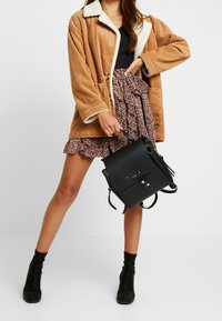 Schwarzer Rucksack mit Klappe, metallischen Beschlägen, kombiniert mit einer braunen Cordjacke und einem gemusterten Rüschensrock. Schwarze Stiefeletten runden das Outfit ab.