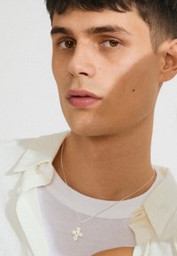 Sølv halskæde med et blomstrets kors vedhæng, båret over en hvid T-shirt, med en lyse farvet knap-op skjorte synlig i baggrunden.
