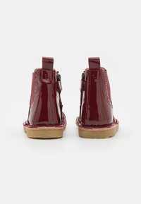Botines de charol rojos con cremalleras laterales y tiradores, que cuentan con una suela plana color marrón claro y un acabado suave y brillante.