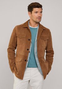 Chaqueta de ante marrón con cuatro bolsillos, cierre de botones y una textura mate. Llevada sobre un suéter azul verdoso y una camisa de cuadros.
