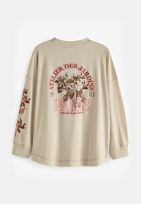 Beige hosszú ujjú pulóver, amely virágmotívummal díszített bordó és rózsaszín színekkel, "PARIS" és "ATELIER DES JARDINS 1983" dekoratív felirattal.