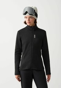 Crna zip-up jakna s visokim ovratnikom, izrađena od glatkog materijala. Nosena uz bijelu rebrastu beanie i velike crne skijaške naočale.