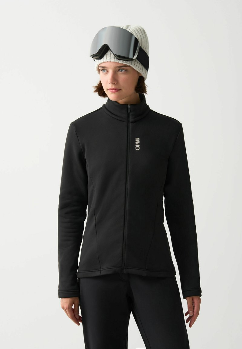 Crna zip-up jakna s visokim ovratnikom, izrađena od glatkog materijala. Nosena uz bijelu rebrastu beanie i velike crne skijaške naočale.