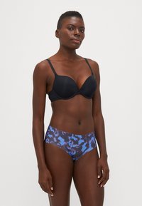 Schwarzer gepolsterter BH mit verstellbaren Trägern, kombiniert mit hochgeschnittenen Unterhosen in einem blauen und lila abstrakten Muster. Glatte Stoffoberfläche.