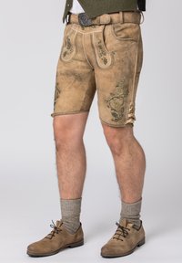 Shorts en cuir marron avec une broderie florale verte, rehaussés de coutures décoratives et de lacets, associés à des chaussures marron et des chaussettes grises.