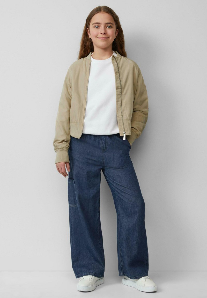 Veste bomber beige, T-shirt blanc, pantalon en denim noir à jambes larges, et baskets blanches. Tissu lisse et coupe décontractée.