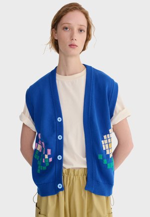 Mladý človek v jasne modrej zapínacej veste s pixelovými kvetinovými vzormi cez bielu košeľu s vyhrnutými rukávmi a béžové nohavice.