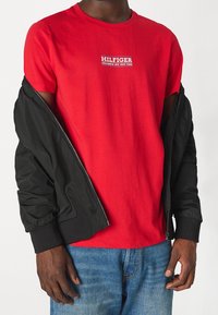 T-shirt di cotone rossa con branding "HILFIGER" in bianco e blu, indossata sotto una giacca bomber nera. Jeans di denim con lavaggio chiaro.