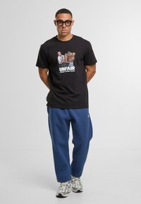 Zwarte katoenen t-shirt met een grafische print met de tekst "UNFAIR" en blauwe joggingsbroek. Model draagt zilveren en grijze sneakers.
