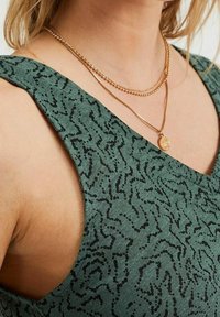 Vihreä hihaton paita, jossa on teksturoitu, aallokkomainen musta kuvio. Sen kaula-aukko on leveä ja siinä on huomaamattomat sivuleikkaukset, jotka on yhdistetty kerroksellisiin kultaketjuihin.