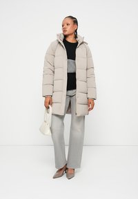 Pufová bunda v svetlo béžovej farbe, s kapucňou, gombíkovým zapínaním a prešívaným dizajnom. Nosená cez šedý sveter, doplnená o šedé džínsy a vysoké podpätky.
