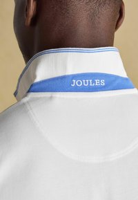 Balta polo krekla ar zilu apkakli, kurā ir teksts "JOULES" baltā krāsā. Apkakles malā ir svītru raksts.