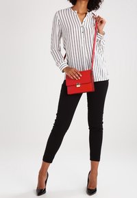 Chemise blanche à manches longues avec des rayures verticales noires, associée à un pantalon noir slim et un petit sac bandoulière rouge. Des talons hauts noirs complètent la tenue.