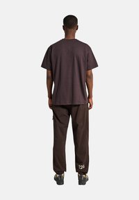 Ciemnobrązowa bawełniana koszulka o luźnym kroju i krótkich rękawach, zestawiona z dopasowanymi brązowymi spodniami z białą grafiką "223" blisko dołu.