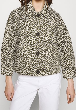 Femme portant une veste courte vert olive avec imprimé floral blanc et gros boutons foncés, associée à un pantalon blanc.