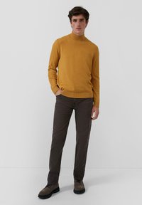 Muž stojí, oblečený v horčicovom roláku, tmavohnedých nohaviciach a hnedých šnurovacích topánkach, s jednou rukou vo vrecku na jednobarevnom pozadí.