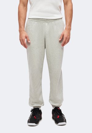 Man wearing light gray sweatpants with elastic cuffs and black sports shoes with red details, standing against a plain background.