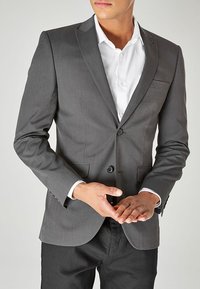 Grau maßgeschneiderte Anzugjacke mit zwei Knöpfen, Reverskragen und Paspeltaschen. Getragen über einem weißen Hemd, das ein klassisches Design hervorhebt.
