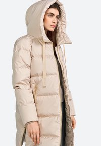 Beige gewatteerde lange jas met een capuchon, voorzien van een ritssluiting en gouden hardware, met een gladde textuur en een horizontaal gestikt patroon.
