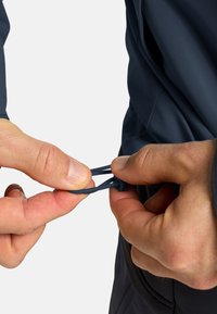 Giacca funzionale blu marina con una texture liscia. Le mani stanno regolando un cordino di chiusura, dotato di un robusto fermacorda in plastica. Dettagli di cucitura sottili visibili.