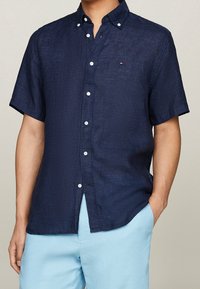 Navy blue short-sleeve button-up shirt with a textured fabric. Features white buttons and a small logo on the chest. Tailored fit with side slits.