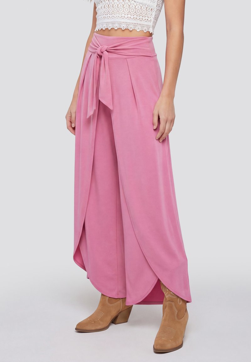 Frau trägt hoch taillierte, rosa, weit geschnittene Hosen mit vorderem Wickeldetail und gebundener Schleife, kombiniert mit beigen Stiefeletten und weißem Spitzen-Crop-Top.
