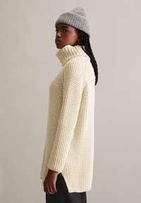 Cremefarbener, übergroßer Strickpullover mit hohem Kragen, strukturiertem Muster, seitlichen Schlitzen und langen Ärmeln, kombiniert mit einer grauen Mütze.