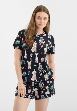 Mujer con blusa de manga corta azul marino y pantalones cortos con estampado multicolor de perros y tenis sobre fondo blanco.