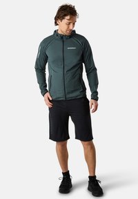 Giacca leggera di colore verde scuro con zip e cappuccio, design elegante e dettagli riflettenti, abbinata a pantaloni corti neri e scarpe sportive.