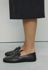 Legs wearing black leather loafers and a black skirt standing on a light gray floor against a white wall.