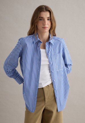 Joven mujer con cabello castaño claro y ondulado lleva una camisa de rayas azules y blancas abotonada sobre una camiseta blanca sin mangas y pantalones beige, de pie con las manos detrás de la espalda.